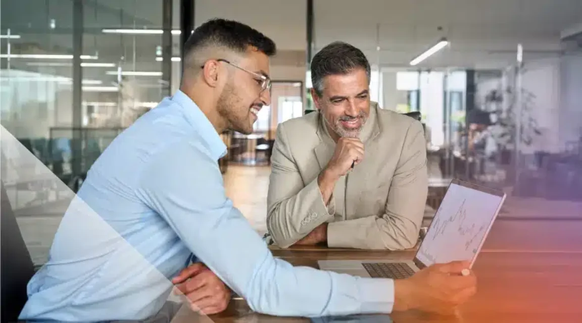 Two professionals review charts and data together on a laptop during a meeting in a modern office.