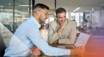 Two professionals review charts and data together on a laptop during a meeting in a modern office.