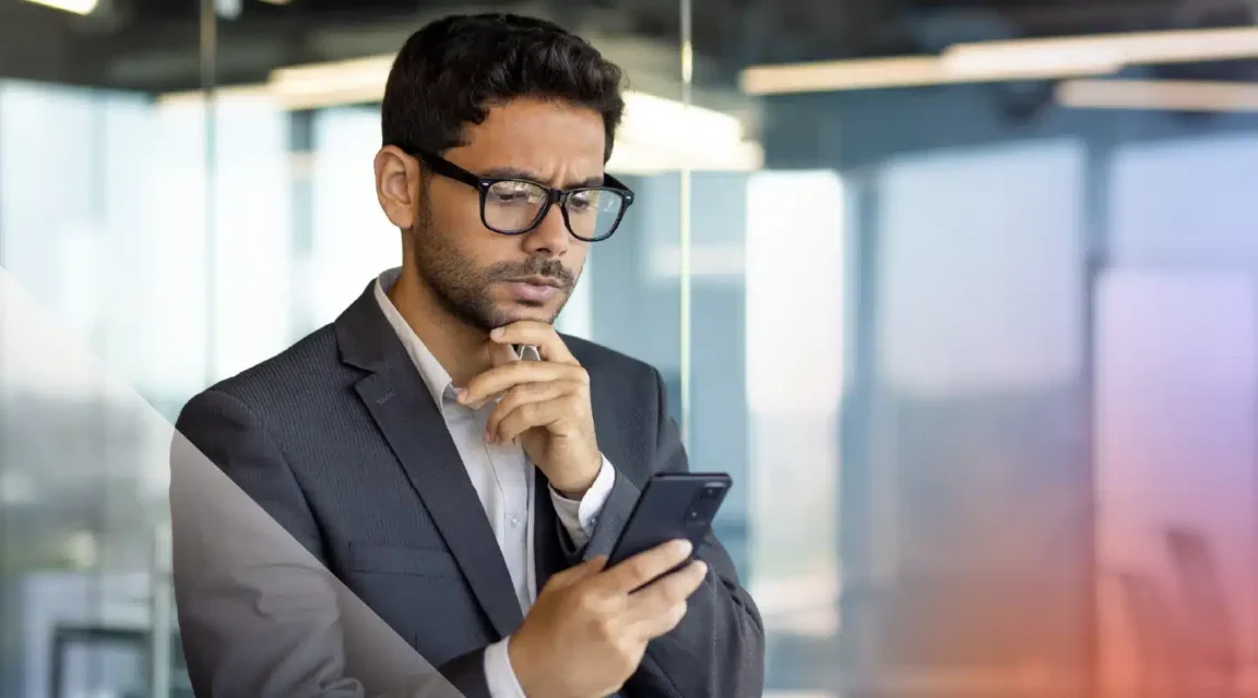 Man in a suit and glasses reading his phone with a concerned expression in a modern glass office.